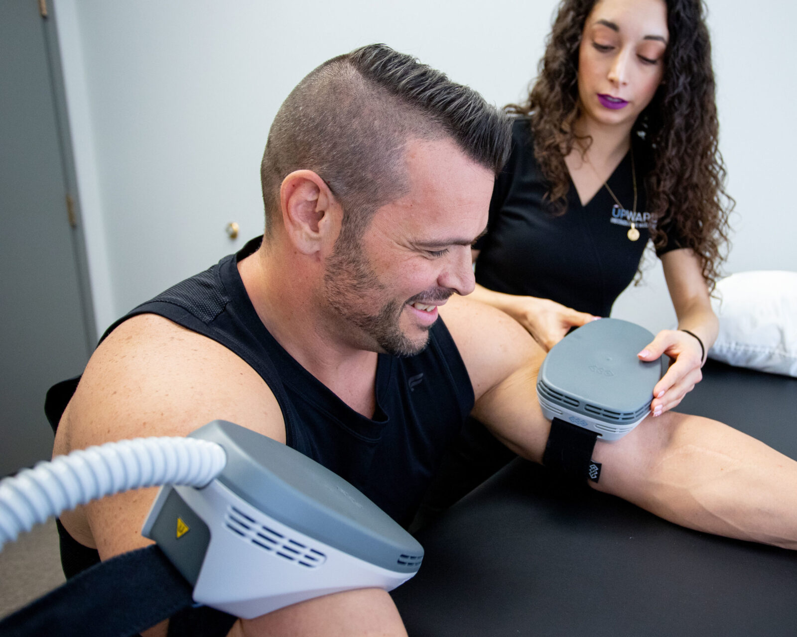 male patient with emsculpt neo body sculpting device on each bicep being aided by female medical professional